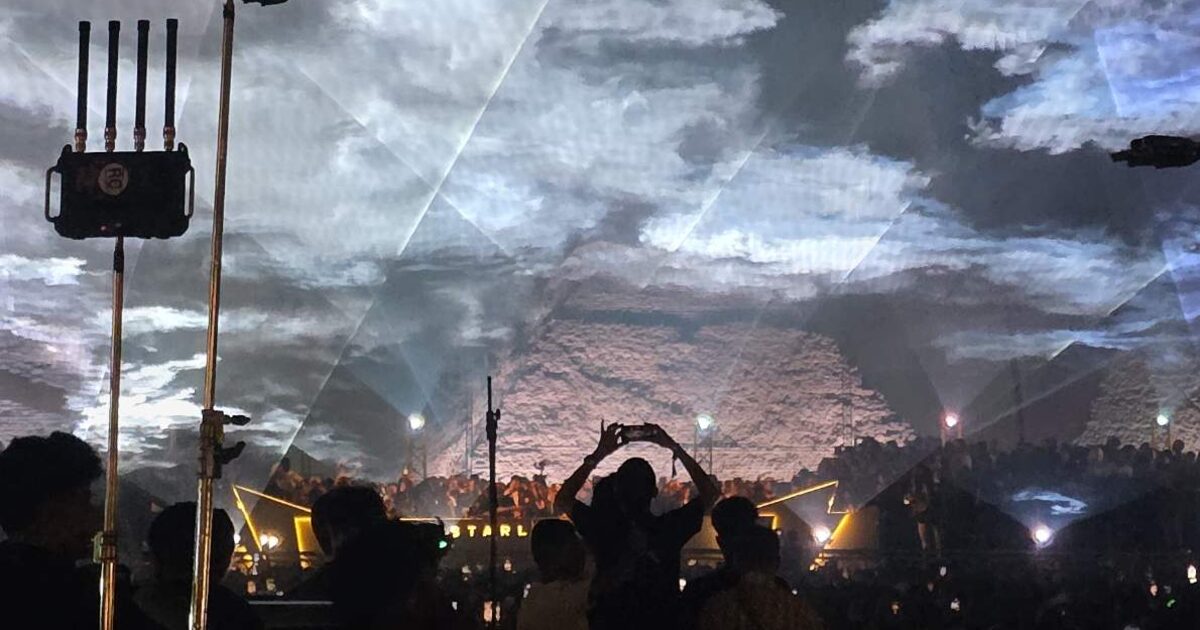 Lasers riscando a nuvem baixa sobre a multidão do Starlight Festival, com a face da pirâmide de Khufu iluminada ao fundo